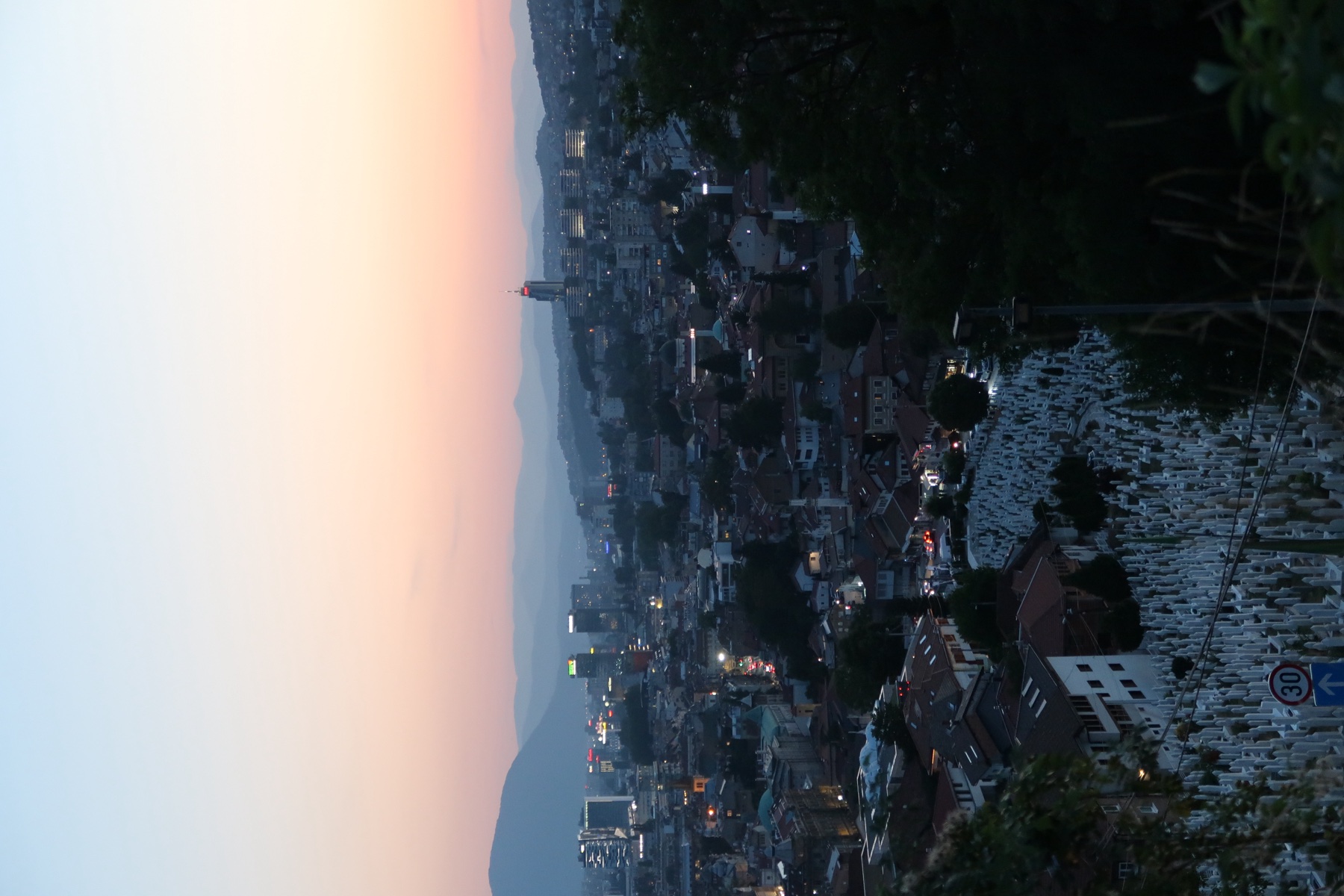 Sarajevo city lights and mountains under a pale orange dusk sky, with white grave markers on the hillside below